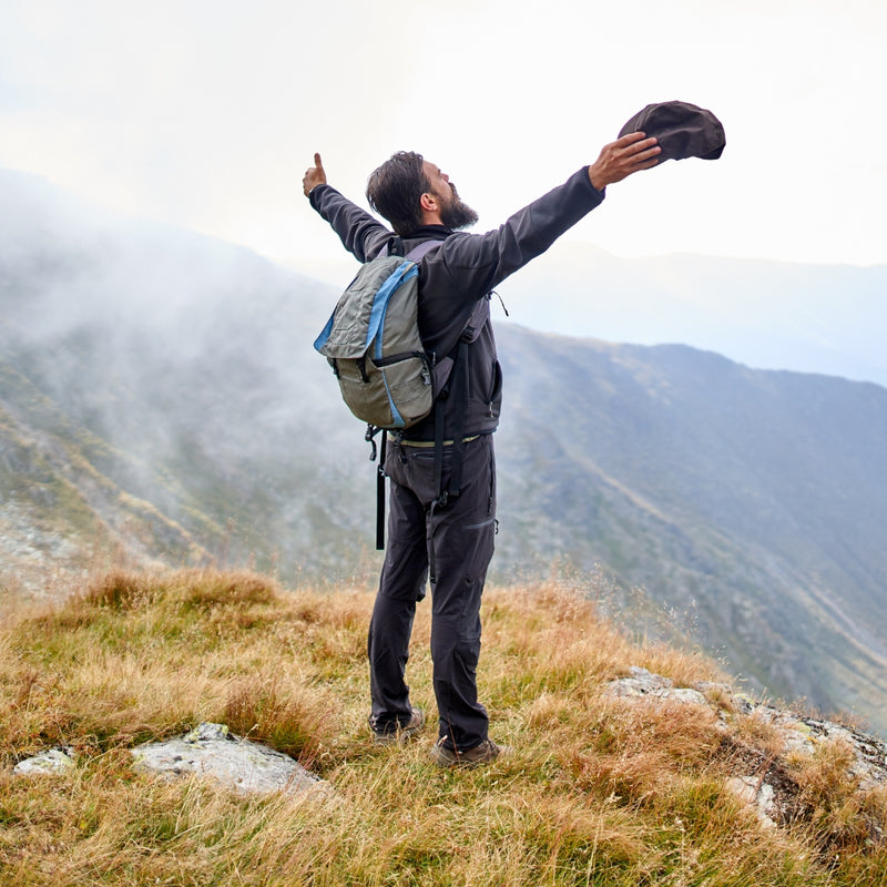 A bearded man in hiking gear stands on a mountain ridge with his arms raised, holding a dark cap above his head. He wears a gray and blue backpack, dark hiking pants, and a dark jacket, with misty mountains in the background.
