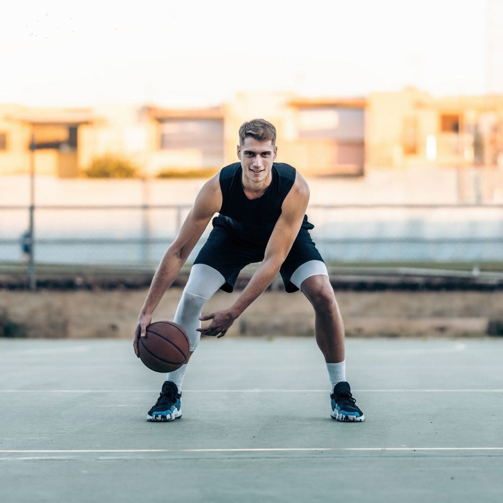 A man wearing a black sleeveless athletic shirt, black shorts, white compression sleeves, and black sneakers dribbling a basketball on an outdoor court.