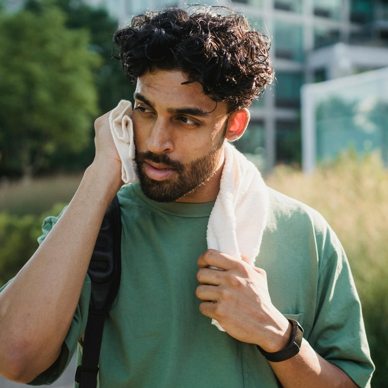 A man outdoors wearing a green t-shirt and holding a white towel to his face, with a black backpack strap visible over his shoulder.