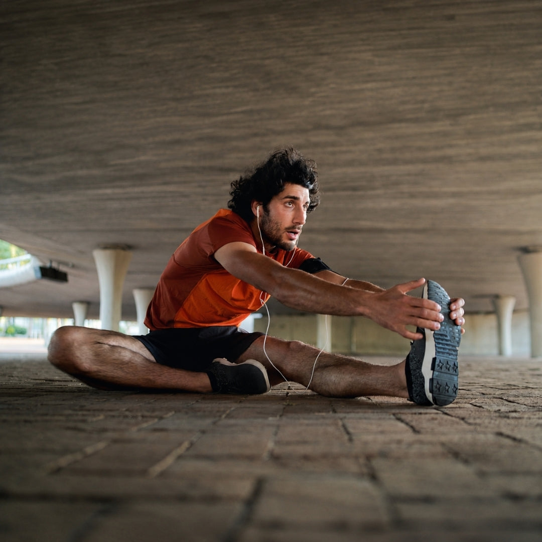 A man in an orange athletic shirt and black shorts stretches his leg on a stone floor under a concrete bridge.