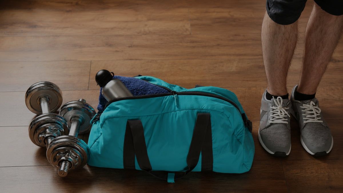 A teal blue duffel bag with black straps sits on a wooden floor next to two dumbbells and a person's legs wearing gray sneakers.