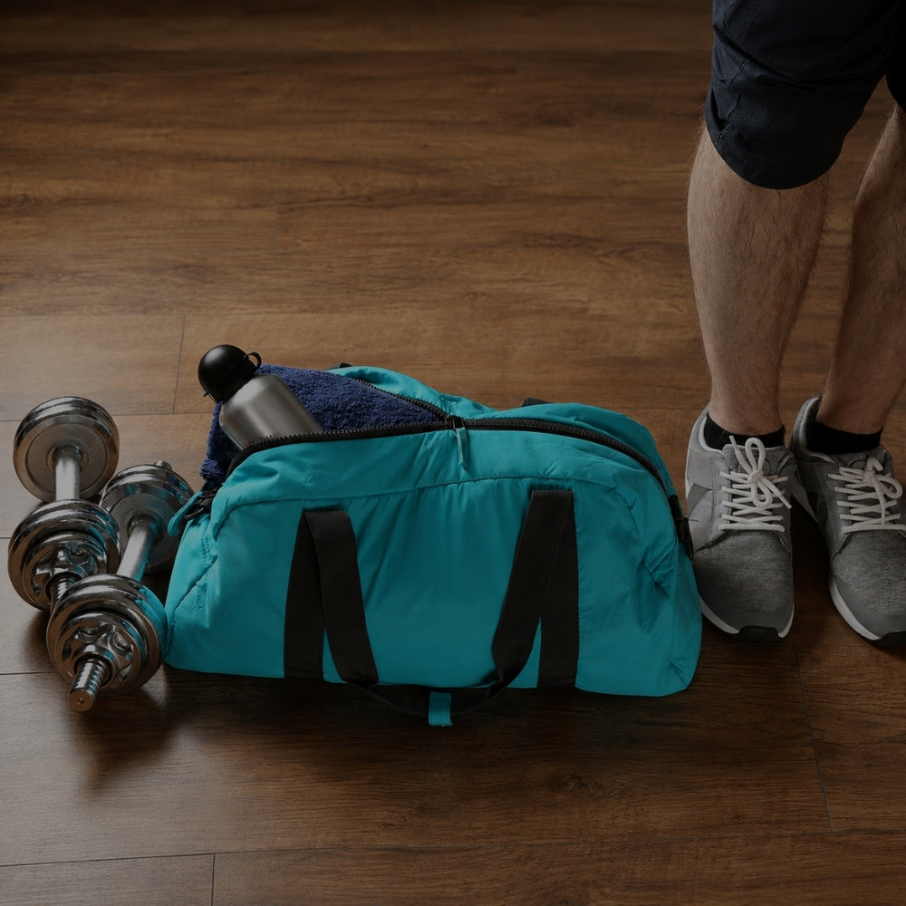A teal duffel bag with black handles sits on a wooden floor next to two silver dumbbells and a person's legs wearing gray sneakers.