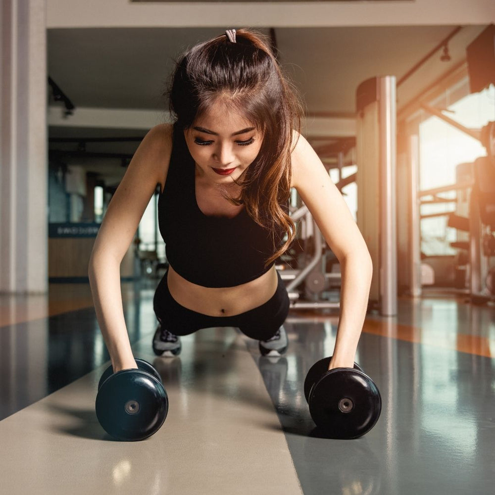 A woman in a black sports bra and leggings does a push-up position on a gym floor while holding two dumbbells, with warm sunlight streaming into the background.