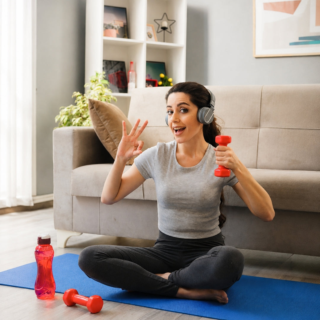 A woman sits cross-legged on a blue exercise mat in a living room, wearing gray headphones and holding a small red dumbbell. She is wearing a light gray t-shirt and black leggings.